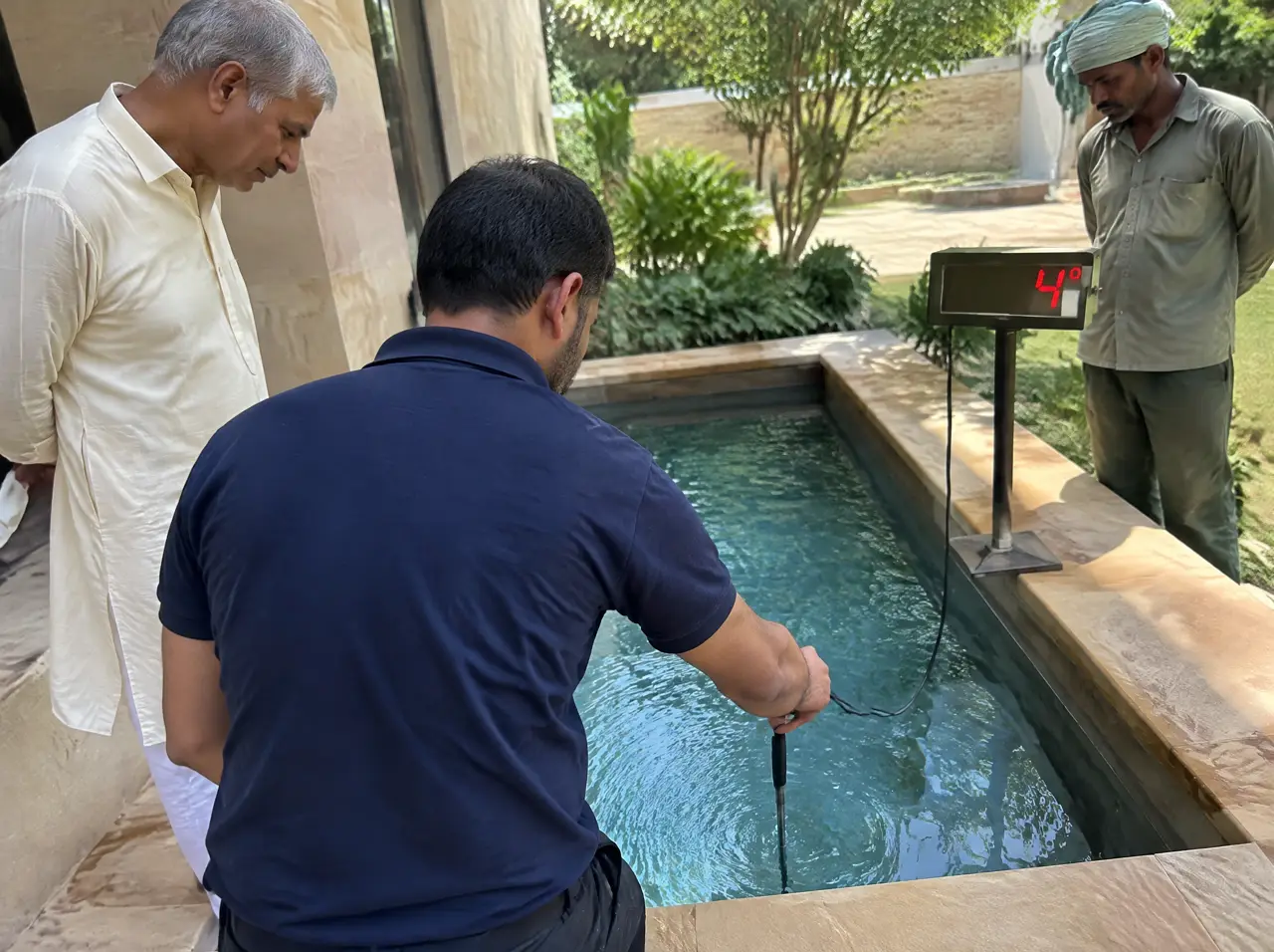 Site engineer dipping a probe thermometer into a cold plunge pool on commissioning day while the owner and gardener watch