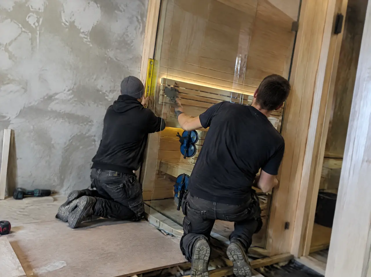 Two installers fitting a frameless tempered-glass panel into a sauna frame with suction lifters