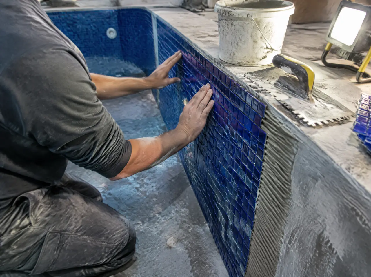 Tiler pressing deep-blue glass mosaic onto the wall of an empty plunge-pool shell